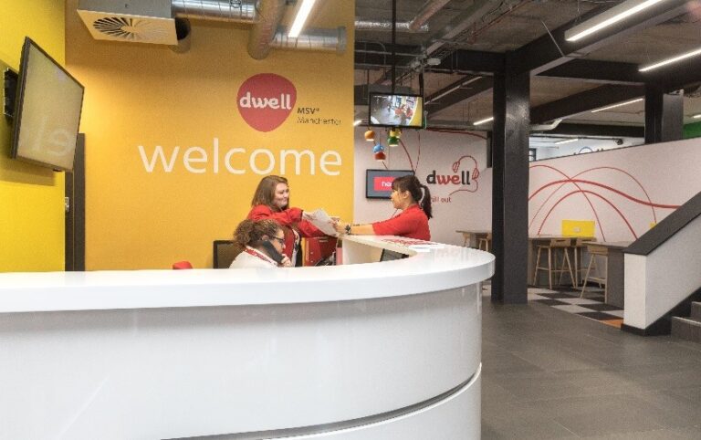 Drei Mitarbeiter in roten Uniformen helfen einem Besucher am weißen Rezeptionstresen einer hell erleuchteten Lobby eines Studentenwohnheims in Manchester mit gelb-weißen Wänden.