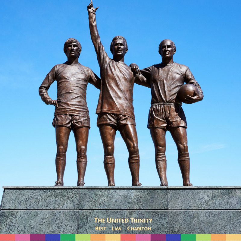Bronze statue of three Manchester football legends standing together on a pedestal, with one holding a ball and another raising an arm; labeled "The United Trinity.