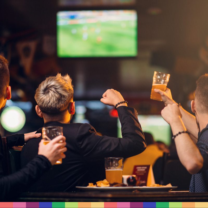 People sitting at a bar in Manchester, holding drinks and cheering while watching an exciting football match on a large TV screen.