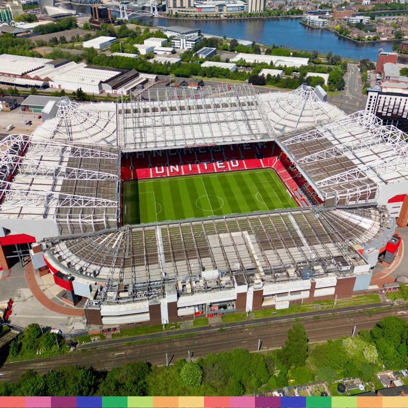 Vista aérea do estádio de futebol Old Trafford, em Manchester, com arquibancadas vazias e um campo de futebol bem conservado, cercado por prédios, estradas e área verde.