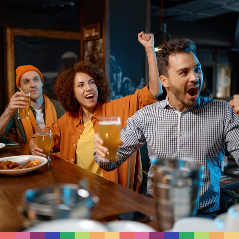 Three people at a bar celebrate while holding beer glasses, appearing excited as they watch a football match off-camera—capturing the lively spirit of Manchester.