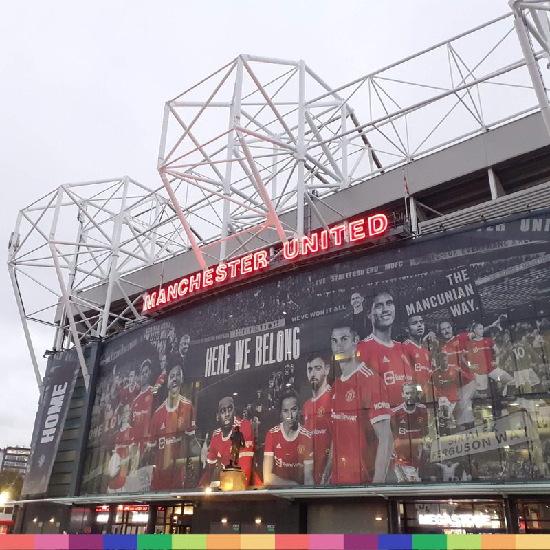 Exterior view of Old Trafford stadium in Manchester, with "Manchester United" in red neon letters above a large mural celebrating the club’s football players.