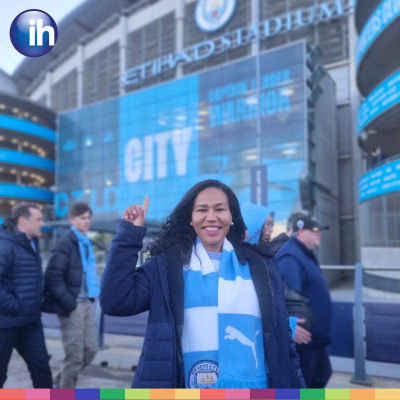 A woman wearing a Manchester City scarf smiles and points upward outside the Etihad Stadium, capturing the vibrant spirit of football in Manchester, with other fans visible in the background.