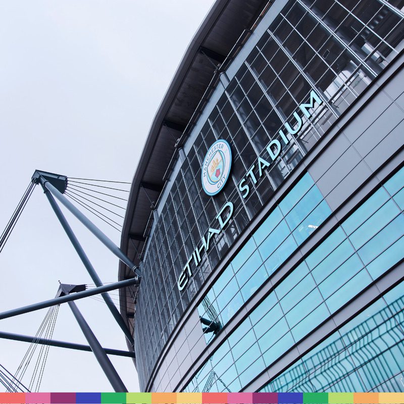 Exterior view of Etihad Stadium in Manchester, featuring large glass windows, the iconic Manchester City Football logo, and "Etihad Stadium" signage visible on the building.