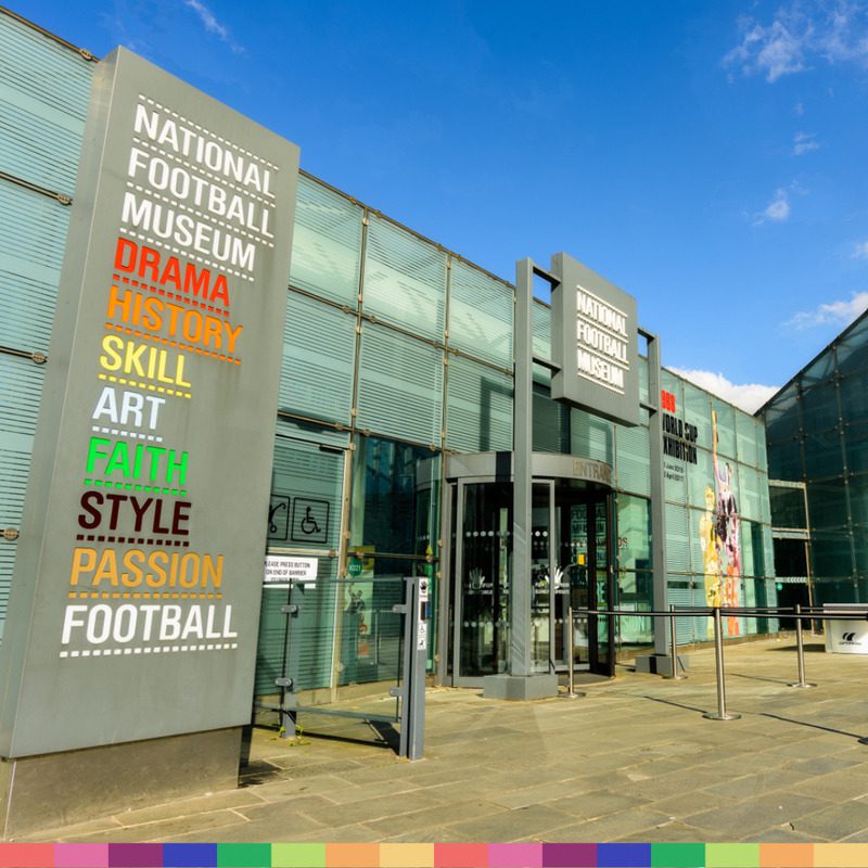 The entrance of the National Football Museum in Manchester, featuring a large sign with words like drama, history, skill, and football, stands out under a clear blue sky.