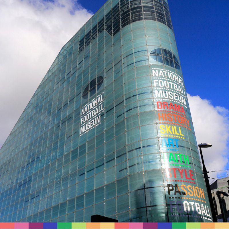 A modern glass building in Manchester labeled "National Football Museum," with colorful vertical text listing qualities like drama, history, skill, and passion.