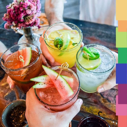 Cuatro manos sostienen coloridos cócteles adornados con rodajas de fruta y hierbas, capturando el animado ambiente de la noche de Manchester. Al fondo se ven un jarrón de flores y una barra de colores.