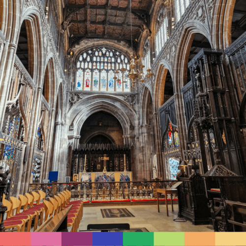 Una vista dell'interno della Cattedrale di Manchester con alti archi in pietra, vetrate colorate, soffitto in legno e file di sedie vuote.