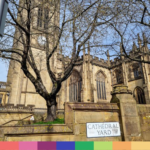 Una grande cattedrale in pietra di Manchester si erge dietro un albero spoglio e un cartello con la scritta "Cathedral Yard" in una giornata di sole.
