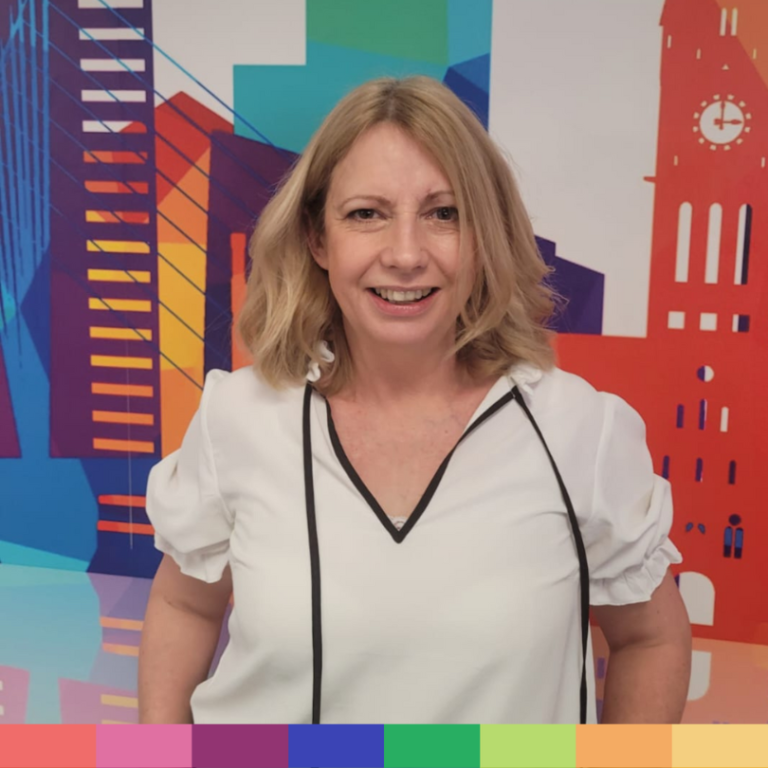 肩までのブロンドの髪に白いブラウスを着た女性が、色鮮やかで抽象的な都市景観の壁画の前に立っており、当社のストーリーの背景にある創造性と精神を体現しています。