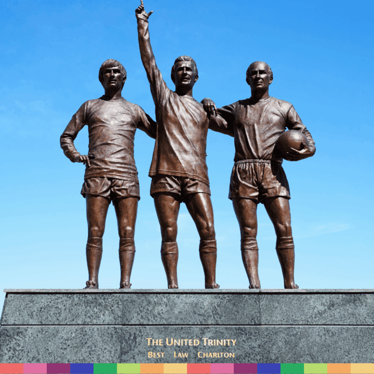 Bronzestatue von drei nebeneinander stehenden Fußballspielern aus Manchester, einer zeigt nach oben, mit der Aufschrift „The United Trinity“ auf dem Sockel vor einem klaren blauen Himmel.