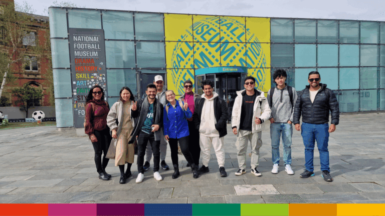 A group of ten people stand and pose for a photo outside the entrance of Manchester's National Football Museum, with signage and modern glass walls visible in the background.