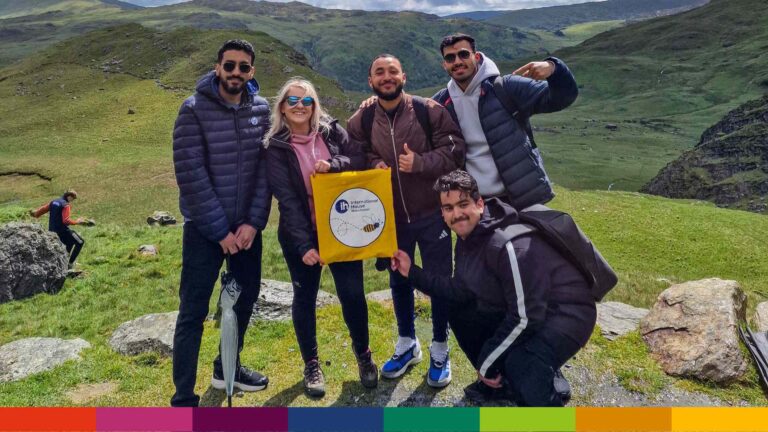 Five people pose outdoors on grassy hills near Manchester, holding a yellow banner. Mountains are visible in the background under a cloudy sky.