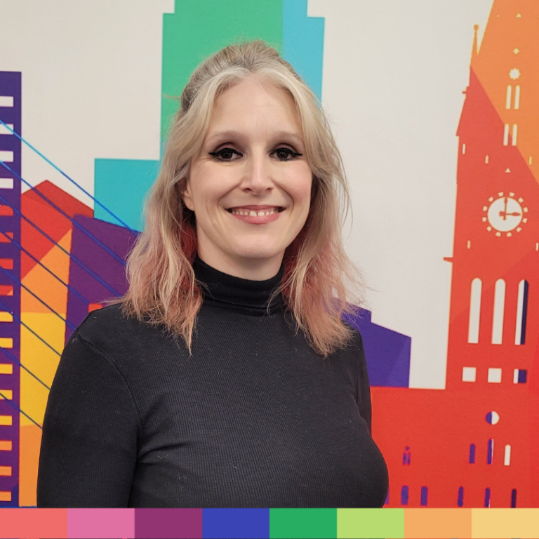 長いブロンドの髪と黒いタートルネックを着た人物が、色鮮やかで抽象的な都市景観の壁画の前で微笑んで立っており、当社の会社概要の活気ある精神を捉えています。