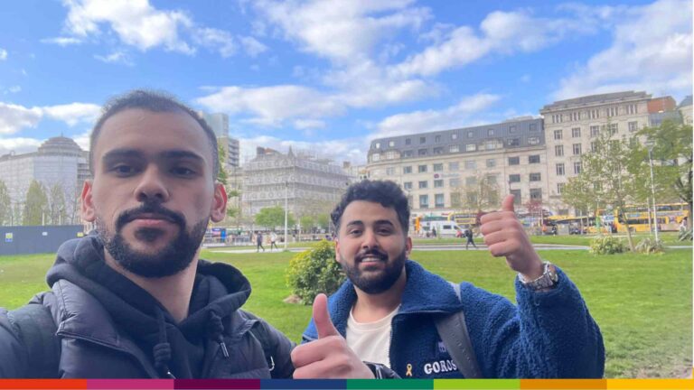 Two men standing outdoors in a Manchester city square on a partly cloudy day, both giving a thumbs-up with buildings and trees visible in the background.