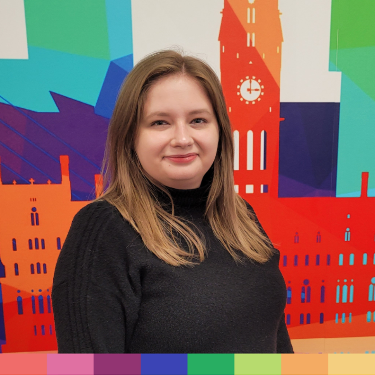 明るい茶色のストレートヘアに黒いセーターを着た女性が、カラフルなイラストが描かれた都市景観の背景の前に立っており、当社の「会社概要」ページの創造的な精神を完璧に表現しています。