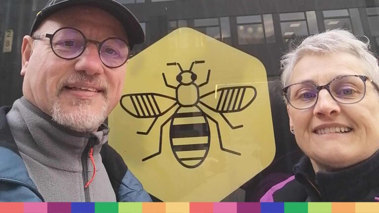 Dos personas con gafas están paradas frente a una ventana con un gran hexágono amarillo que representa una abeja negra, que simboliza Manchester, una ciudad conocida por recibir a estudiantes internacionales y ofrecer oportunidades de estudiar en el extranjero.