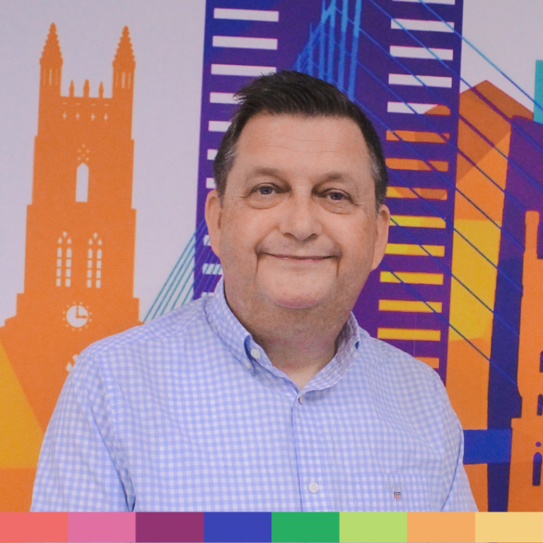 薄紫色のチェック柄シャツを着た男性が、建物や橋を描いたカラフルな都市景観を描いた背景の前に立っており、当社の「会社概要」ページの歓迎の精神を完璧に表現しています。