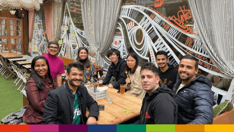 A group of nine people sit around a wooden table at an outdoor cafe in Manchester, smiling at the camera, with patterned curtains and murals in the background.