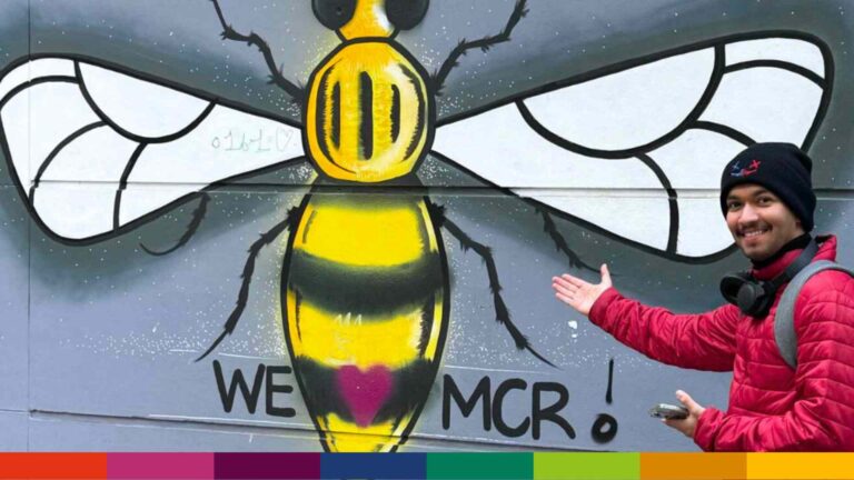 A person in a red jacket points at a mural of a bee with the text "WE [heart] MCR!" on a wall, celebrating Manchester’s vibrant spirit.