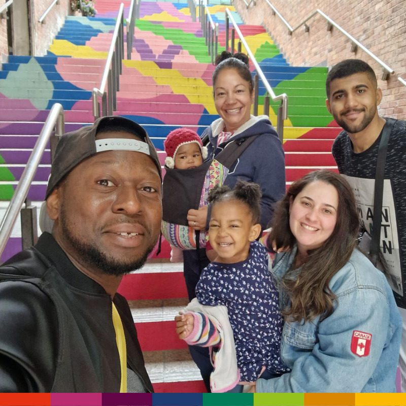 A group of five people, including two young children and their friendly homestay host, smile at the camera in front of brightly painted, colorful stairs.
