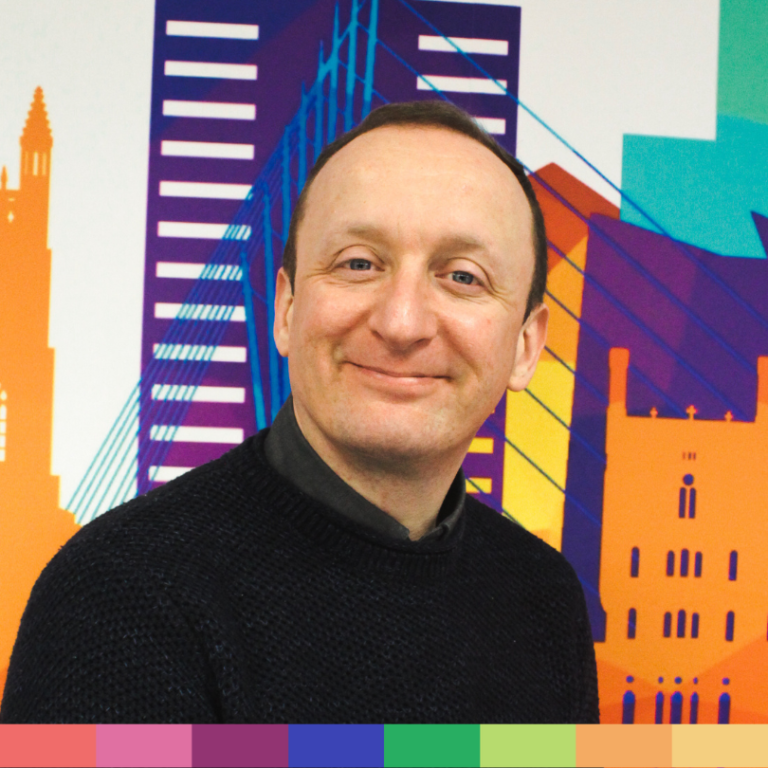 ショートヘアの男性が、カラフルな街並みを描いたイラストを背景に、カメラに向かって微笑んでいます。下部には色とりどりの長方形が並び、この活気あふれる「About Us」イメージに親しみやすさとクリエイティブな雰囲気を与えています。