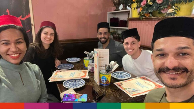 Five people wearing traditional hats sit around a restaurant table in Manchester, smiling at the camera, with menus, dishes, and floral decor in the background.