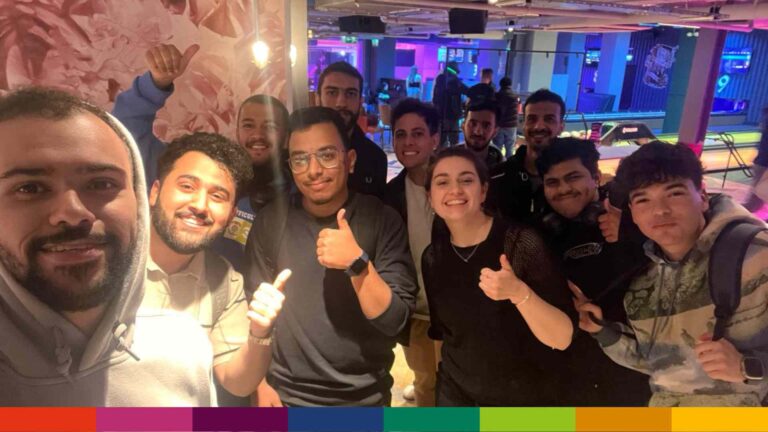 A group of people stand indoors in Manchester, smiling and giving thumbs up to the camera. Pool tables and neon lights are visible in the background.