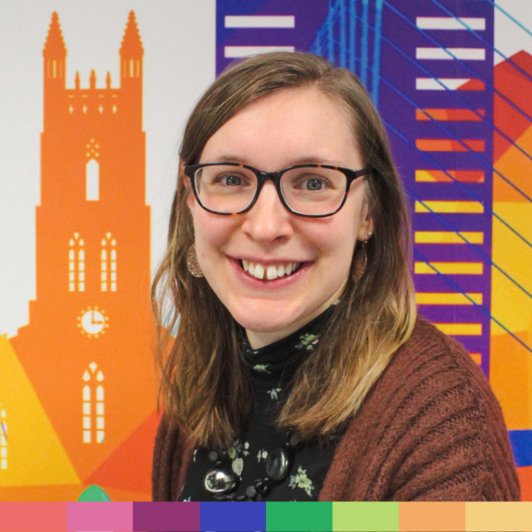 眼鏡をかけ、肩までの長さの髪をかけた女性が、オレンジ色の時計塔を描いたカラフルな街並みの壁画の前で微笑んでおり、当社の「会社概要」ページのフレンドリーな雰囲気を完璧に表現しています。