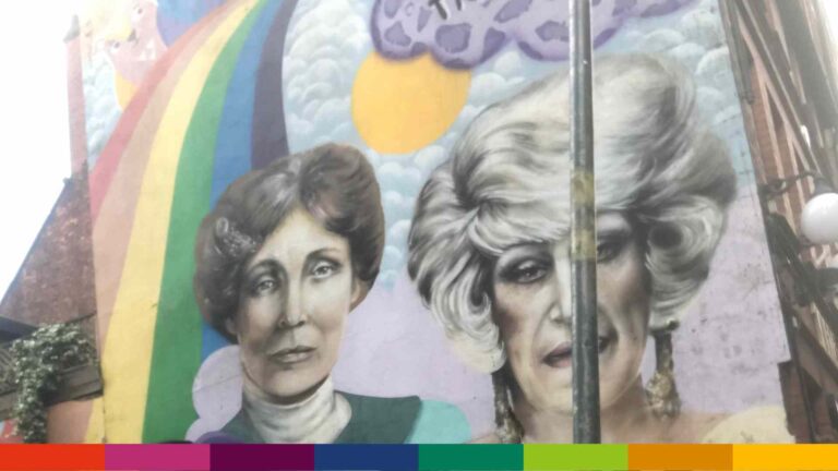 A mural in Manchester featuring two women’s faces with a rainbow and clouds in the background, partially obscured by a lamppost.
