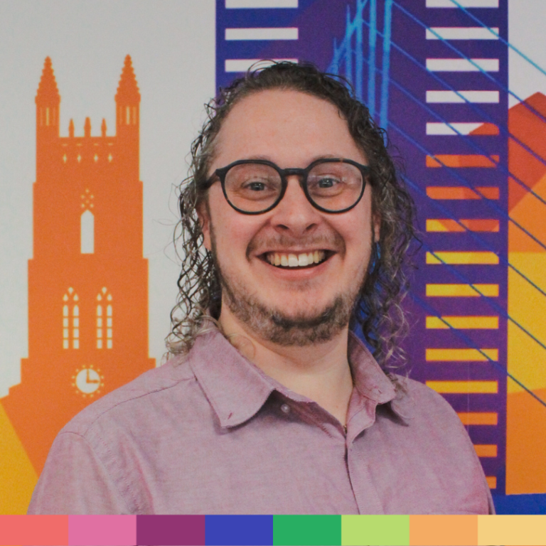 巻き毛、眼鏡、薄紫色のシャツを着た人物が、カラフルな幾何学模様の街並みを描いた壁画の前で微笑んでいます。魅力的な「会社概要」ページにぴったりです。画像の下部には、カラーブロックの列が見えます。