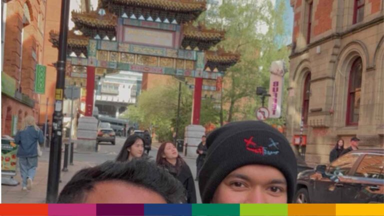 Two people pose for a selfie in front of a traditional Chinese archway on a Manchester city street, with other pedestrians and cars in the background.
