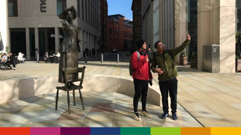 Two people stand next to a bronze statue on a Manchester sidewalk; one wears a red jacket, the other takes a selfie with a phone. Buildings and outdoor seating are visible in the background.