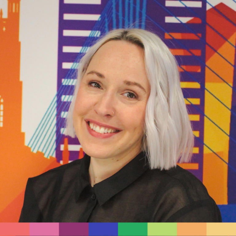 プラチナブロンドのショートヘアと黒いシャツを着た人物が、カラフルな幾何学的な都市景観の壁画の前で微笑んでおり、当社のストーリーの活気ある精神を反映しています。