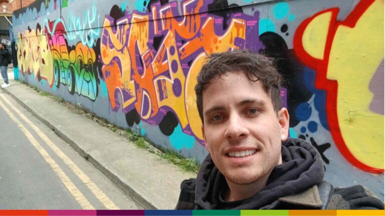 A man takes a selfie on a Manchester street with colorful graffiti art covering a wall behind him.