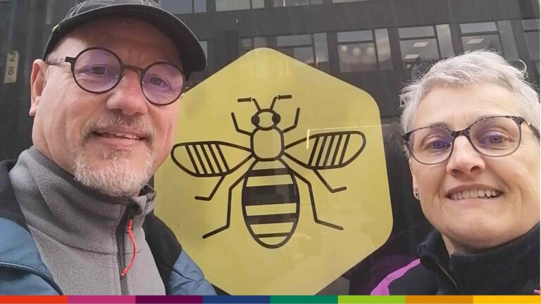 Two people wearing glasses pose in front of a window with a large yellow hexagon featuring Manchester’s iconic black bee design.