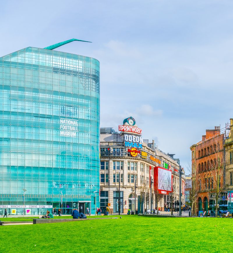 Un edificio in vetro con la scritta "National Football Museum" sorge accanto a edifici storici e moderni, nei pressi di un prato verde nel centro di Manchester.