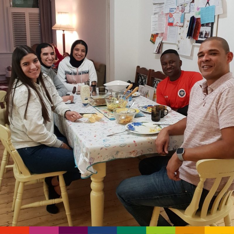 Five people, including their friendly homestay host, are sitting around a dining table, smiling at the camera. The table is set with food, drinks, and utensils in a cozy, well-lit room.