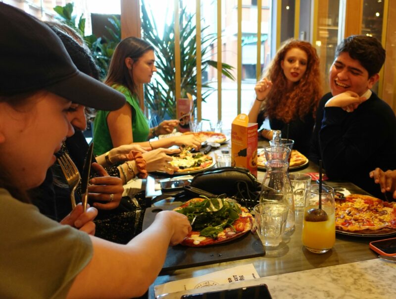 Um grupo de pessoas está sentado ao redor de uma mesa em um restaurante, saboreando pizza e bebidas enquanto conversam. Pratos e talheres estão espalhados pela mesa enquanto participam de animadas atividades sociais.