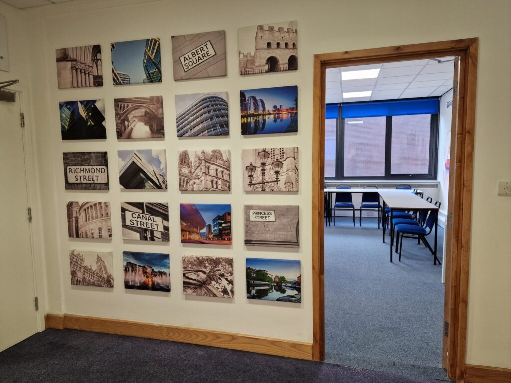 Wall with framed black-and-white city photographs and street signs beside an open doorway leading to a classroom with tables and chairs at IH Manchester English School.
