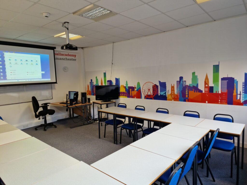 Classroom at IH Manchester English School with U-shaped desk arrangement, blue chairs, a projector screen displaying a computer desktop, and a colorful cityscape mural on the wall.