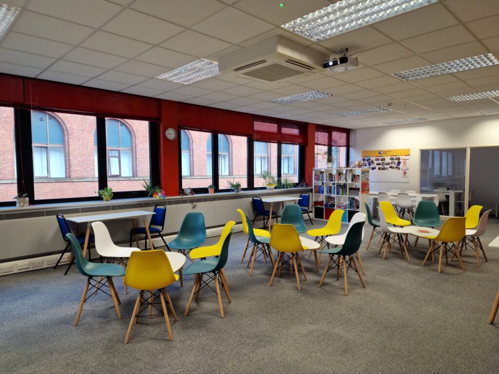 Modern office break room with colorful chairs around tables, large windows, plants on the windowsill, and a bulletin board on the back wall—perfect for students at IH Manchester English School to relax and connect.