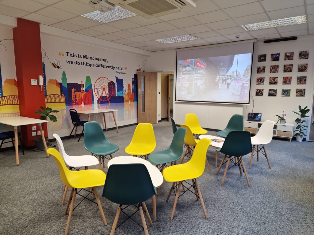 A classroom at IH Manchester English School with colorful chairs arranged in a circle, a projector screen displaying a street scene, vibrant wall art, and various plants and decor.