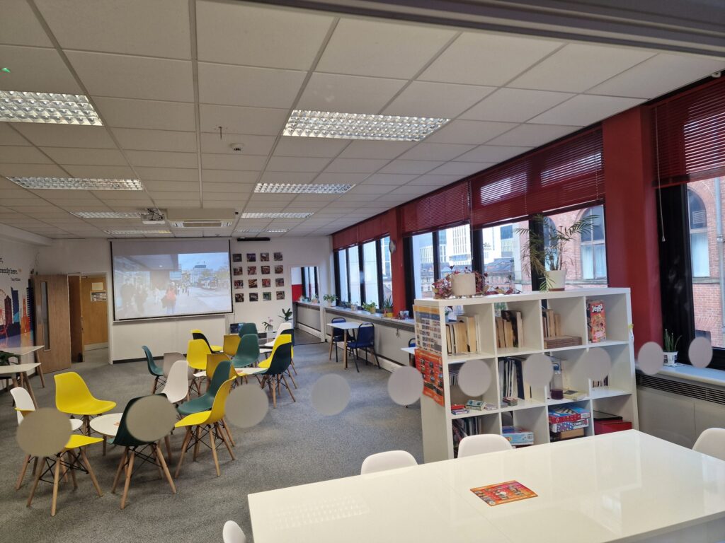 A modern classroom at IH Manchester English School with colorful chairs, round tables, bookshelves, and large windows; a projector displays an image on the front wall.