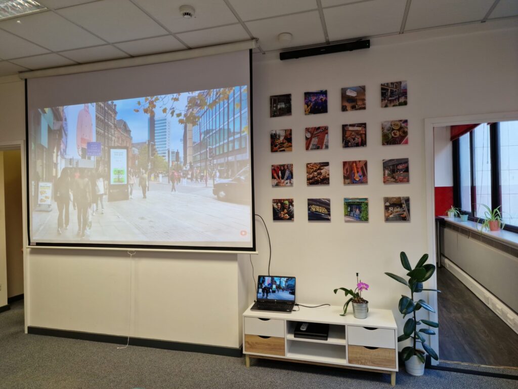 A projector at IH Manchester English School displays a city street scene on a screen in an office, with a laptop, potted plant, and photos decorating the nearby wall.