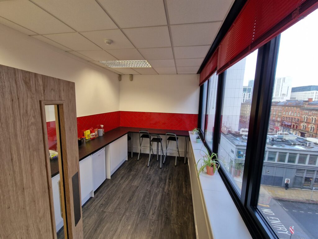 Small office kitchen at IH Manchester English School with white cabinets, black counters, a red backsplash, bar stools, a kettle, and a window overlooking city streets and buildings.