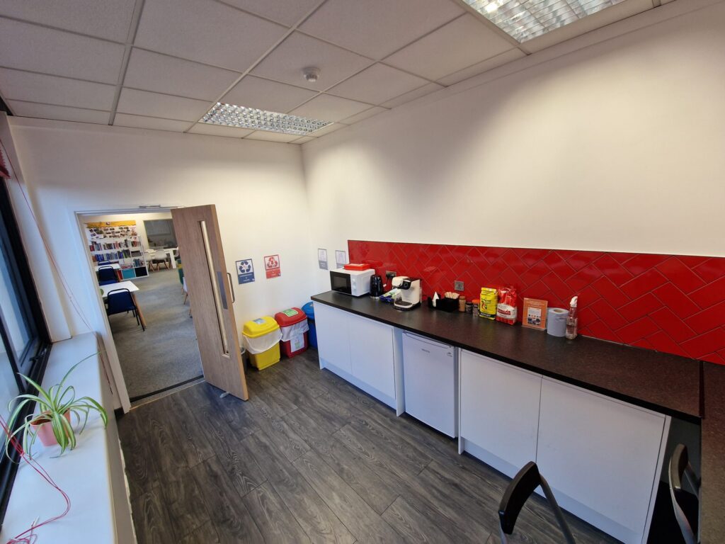 Small office kitchen at IH Manchester English School with a red backsplash, countertop appliances, snacks, bins, and a view into the office area with desks and bookshelves through an open door.
