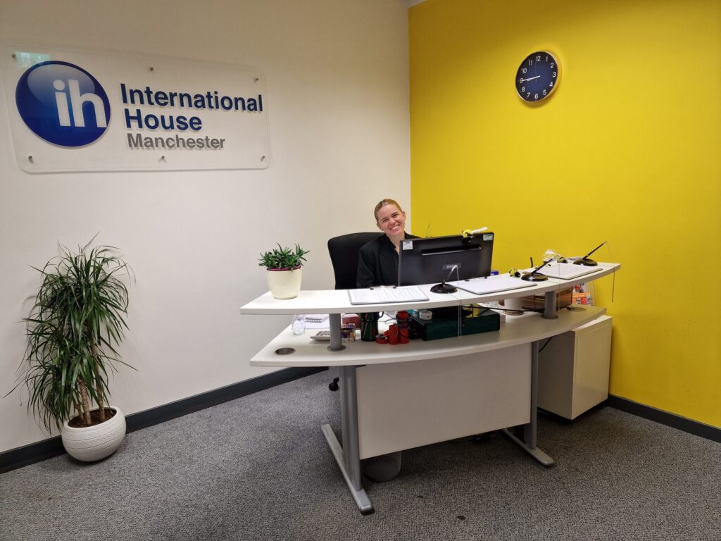 A person sits at the reception desk of IH Manchester English School’s modern office, with “International House Manchester” signage and a yellow accent wall featuring a clock.