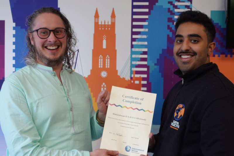 Dos hombres sonríen mientras uno entrega un Certificado de Finalización al otro, celebrando un logro arraigado en la Promesa al Cliente de IH. Al fondo se ve un colorido mural de paisaje urbano.