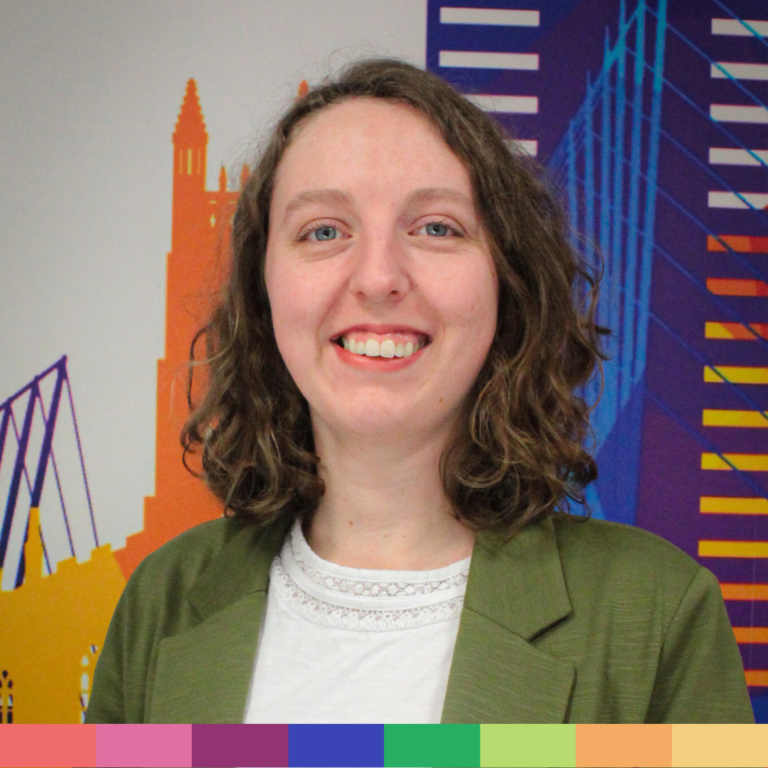 緑色のブレザーと白いトップスを着た茶色の巻き毛の女性が、カラフルで抽象的な都市景観の背景の前に立っており、当社の「会社概要」ページのフレンドリーでプロフェッショナルな精神を体現しています。